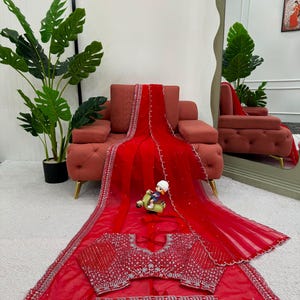 Peut inclure: Un sari rouge vif orn&eacute; de broderies argent&eacute;es complexes et de miroirs le long des bordures et sur le chemisier assorti. Le tissu transparent est agr&eacute;ment&eacute; de petites d&eacute;corations argent&eacute;es &eacute;parpill&eacute;es, formant un ensemble d&eacute;taill&eacute;.