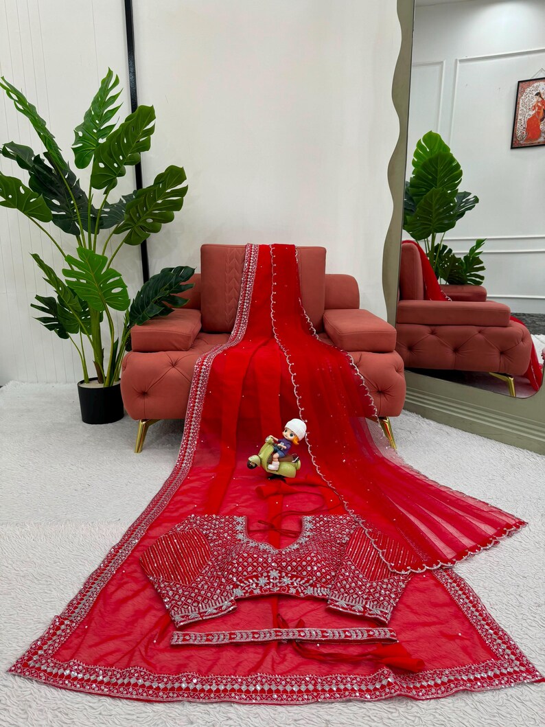 Peut inclure: Un sari rouge vif orn&eacute; de broderies argent&eacute;es complexes et de miroirs le long des bordures et sur le chemisier assorti. Le tissu transparent est agr&eacute;ment&eacute; de petites d&eacute;corations argent&eacute;es &eacute;parpill&eacute;es, formant un ensemble d&eacute;taill&eacute;.