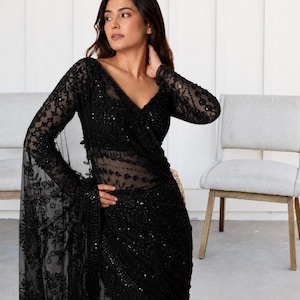 May include: A black long-sleeved saree featuring intricate floral embroidery and sequin embellishments throughout the sheer fabric. The garment has a deep V-neckline and a fitted bodice, with a draped pallu that showcases detailed needlework.