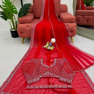 Peut inclure: Un sari en filet rouge vif orn&eacute; de broderies argent&eacute;es complexes et de miroirs le long des bordures. L'ensemble comprend un chemisier rouge assorti avec des ornements argent&eacute;s denses et une ceinture rouge coordonn&eacute;e avec des d&eacute;tails argent&eacute;s.