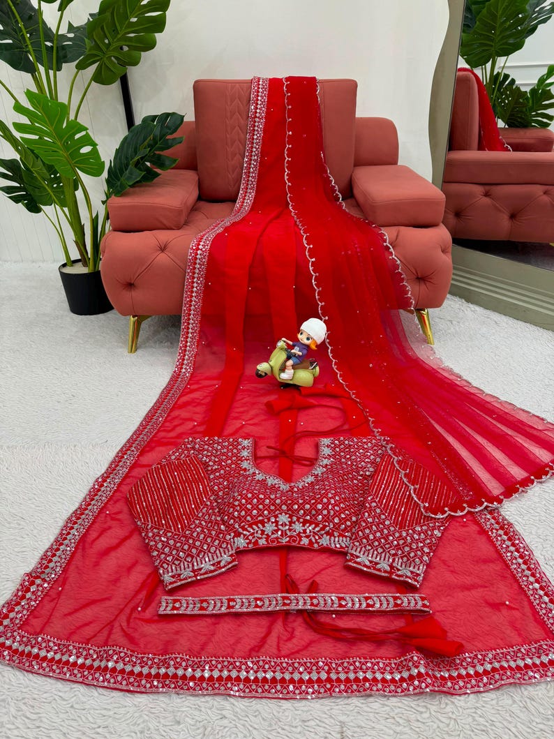 Peut inclure: Un sari en filet rouge vif orn&eacute; de broderies argent&eacute;es complexes et de miroirs le long des bordures. L'ensemble comprend un chemisier rouge assorti avec des ornements argent&eacute;s denses et une ceinture rouge coordonn&eacute;e avec des d&eacute;tails argent&eacute;s.