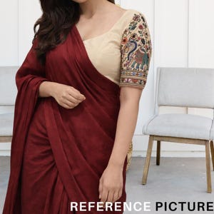 Könnte beinhalten: Ein bordeauxfarbener Sari mit einer beigen Bluse. Die Bluse hat bestickte Ärmel mit Blumen- und Pfauenmotiven in verschiedenen Farben. Der Text "REFERENCE PICTURE" befindet sich unten.