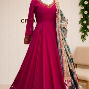 Puede incluir: Un vestido largo de color magenta con mangas largas, con bordados dorados alrededor del cuello redondo y los puños. Un chal estampado con motivos florales en azul verdoso, beige y negro está drapeado sobre un hombro con borde a rayas.