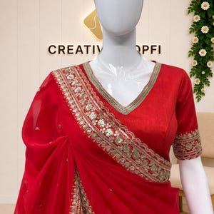 May include: A vibrant red saree featuring a V-neck blouse with short sleeves. The garment is adorned with intricate gold embroidery, sequins, and metallic thread work along the borders and across the sheer fabric, creating a detailed pattern.