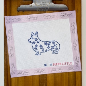 May include: A white needlepoint canvas featuring a blue floral-patterned corgi silhouette in the center. The edges are bound with light purple tape printed with the text Pippa Little. The text Pippa Little is also printed in red near the bottom right.
