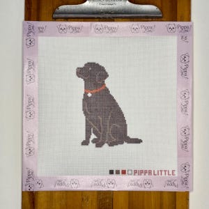 May include: A square needlepoint canvas featuring a dark brown Labrador retriever sitting in the center. The dog wears an orange collar with a white tag. The canvas has a light purple border printed with the text Pippa Little and a dog icon.
