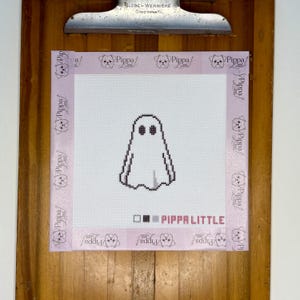May include: A white needlepoint canvas featuring a black outlined ghost design with two oval eyes. The canvas is bordered by light pink tape printed with the text Pippa Little and includes a color key and the text Pippa Little at the bottom.