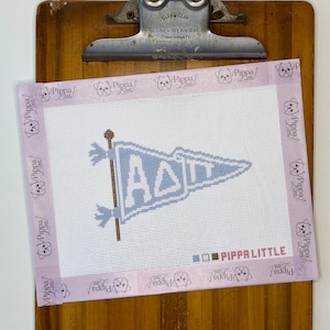 May include: A needlepoint canvas featuring a light blue pennant flag with the Greek letters Alpha Delta Pi in white. The canvas is bordered by light purple tape printed with the text Pippa Little and includes a color key with the text Pippa Little.