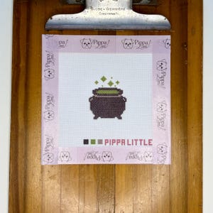 May include: A square needlepoint canvas featuring a dark purple cauldron with a green bubbling liquid and green sparkles above it. The canvas is bordered by light purple tape printed with the text Pippa Little and dog icons, with a color key at the bottom.
