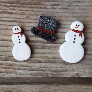 May include: Wooden snowman ornaments with black button eyes, a red scarf, and a black top hat with a red band. The snowman has a white body. The hat is black with white speckles. The ornaments are on a wooden surface.