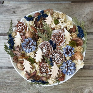 May include: A white ceramic bowl filled with a decorative potpourri arrangement. The arrangement includes brown pine cones, blue-painted pine cones, small wooden tree cutouts, dried berries, and cinnamon sticks. The bowl sits on a wooden surface.