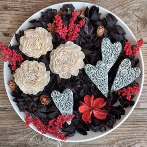 May include: A white circular bowl filled with black potpourri, three cream-colored wooden flowers, three white heart-shaped ornaments with black intricate patterns, small brown pinecones, and sprigs of bright red dried floral accents.