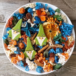 May include: A white bowl filled with colorful potpourri and three green gnome-shaped decorations. The potpourri includes blue, orange, and white dried flowers, cinnamon sticks, and orange slices. The gnomes have white beards and pointed hats.