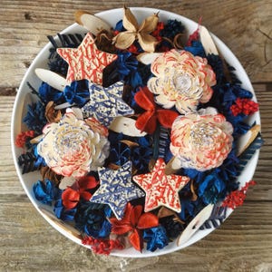 May include: A white ceramic bowl filled with decorative elements in red, white, and blue. The arrangement includes star-shaped ornaments, flower-like decorations, and dried botanicals, creating a patriotic theme.