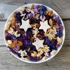 May include: A white ceramic bowl filled with a potpourri arrangement. The potpourri includes dried flowers, star-shaped decorations, and spices in shades of purple, blue, beige, and brown. The bowl sits on a weathered wooden surface.