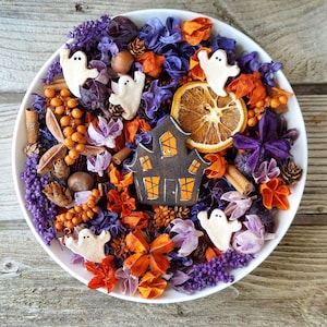 May include: A white bowl filled with Halloween-themed potpourri featuring purple and orange dried flowers, cinnamon sticks, pinecones, hazelnuts, a dried orange slice, and several small white ghost figures surrounding a central dark house cutout.