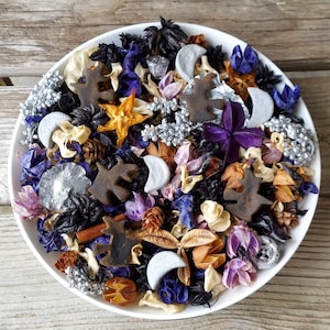 May include: A white ceramic bowl filled with a decorative potpourri arrangement. The potpourri includes dried flowers, leaves, and decorative elements in shades of purple, silver, brown, and orange. The arrangement features star and moon shapes.