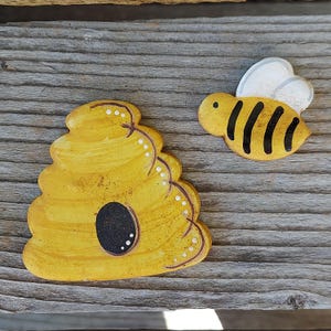 May include: Wooden cutouts of a yellow beehive and a bee. The beehive is yellow with a black opening and white dot accents. The bee is yellow with black stripes and white wings. The items are on a weathered wooden surface.