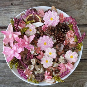 May include: A white bowl filled with a decorative potpourri arrangement. The arrangement includes pink and white artificial flowers, dried orange slices, pine cones, and various dried botanicals. The potpourri is arranged on a wooden surface.
