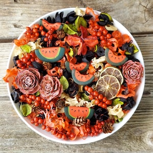May include: A white bowl filled with a colorful potpourri arrangement. The arrangement includes red berries, dried orange slices, watermelon slices, and rose-shaped ornaments. The potpourri has a variety of textures and colors, including orange, red, green, black, and brown.