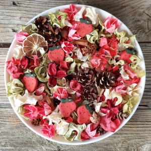 May include: A white bowl filled with a potpourri arrangement. The potpourri includes red, pink, and cream-colored dried flowers, small pine cones, and strawberry-shaped decorations. A dried citrus slice and cinnamon stick are also visible.