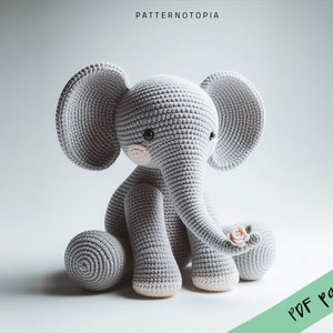 Könnte beinhalten: Ein gehäkeltes graues Elefanten-Amigurumi-Spielzeug mit rosa Akzenten und einer kleinen rosa Blume im Rüssel. Das Spielzeug sitzt auf einer weißen Oberfläche.  PDF-Muster.