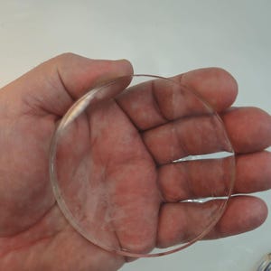 Könnte beinhalten: Eine transparente, kreisförmige Linse, die in einer Hand gehalten wird. Die Linse ist klar und scheint aus Glas oder einem ähnlichen Material zu bestehen. Die Kanten sind glatt und abgerundet. Sie ist wahrscheinlich für Brillen oder ein anderes optisches Gerät.