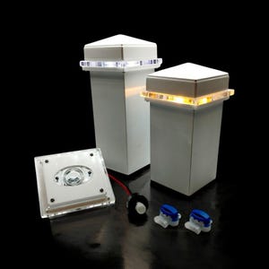 Puede incluir: Dos luces de tapa de poste cuadradas blancas con una cubierta de plástico transparente y una luz LED blanca en el interior. Las luces están conectadas a una fuente de alimentación blanca con un cable negro. Hay dos conectores de cable azules.