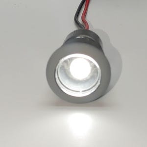Puede incluir: Una pequeña luz LED redonda y gris con una lente blanca. La luz está cableada con cables rojo y negro.