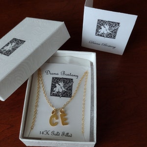 This image shows a Gold initial necklace mounted on a logo card in a Diana Bostany Logo White Swirl Gift Box