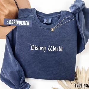 Puede incluir: Una sudadera azul marino bordada con las palabras "Disney World" en escritura blanca. La sudadera tiene mangas largas y cuello redondo. Un collar dorado con un colgante est&aacute; drapeado en la parte delantera. El texto "Embroidered" est&aacute; en una etiqueta azul.