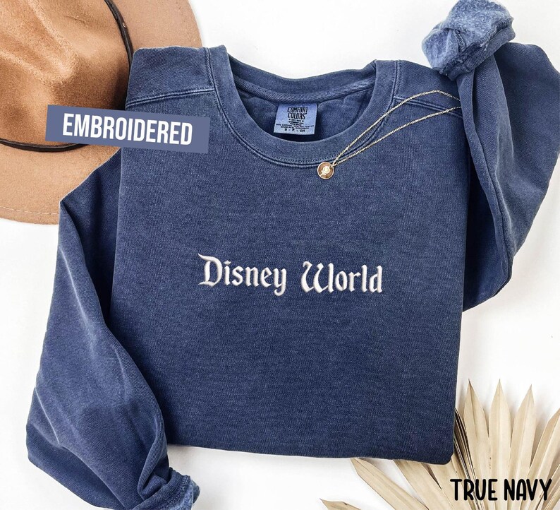 Puede incluir: Una sudadera azul marino bordada con las palabras "Disney World" en escritura blanca. La sudadera tiene mangas largas y cuello redondo. Un collar dorado con un colgante est&aacute; drapeado en la parte delantera. El texto "Embroidered" est&aacute; en una etiqueta azul.