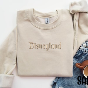 Puede incluir: Una sudadera de color arena con la palabra "Disneyland" bordada en un tono a juego. La sudadera se muestra con vaqueros azules y una taza marr&oacute;n llena de un l&iacute;quido oscuro. La palabra "SAND" est&aacute; impresa en negro.