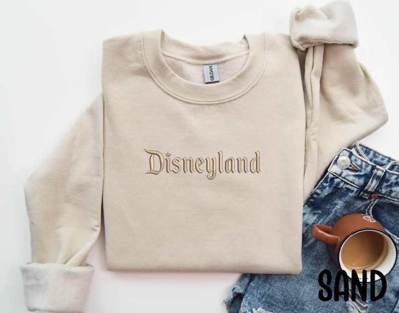 Puede incluir: Una sudadera de color arena con la palabra "Disneyland" bordada en un tono a juego. La sudadera se muestra con vaqueros azules y una taza marr&oacute;n llena de un l&iacute;quido oscuro. La palabra "SAND" est&aacute; impresa en negro.
