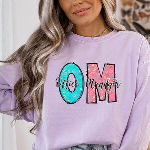May include: A lavender colored long-sleeved sweatshirt with the letters "OM" in a colorful patterned design. The letters stand for "Office Manager" and are written in a playful font.