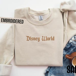 Puede incluir: Sudadera bordada de color arena con el texto "Disney World" en una fuente decorativa. La palabra "EMBROIDERED" est&aacute; impresa en una etiqueta blanca. La sudadera se combina con vaqueros azules y una taza marr&oacute;n.