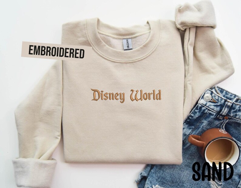 Puede incluir: Sudadera bordada de color arena con el texto "Disney World" en una fuente decorativa. La palabra "EMBROIDERED" est&aacute; impresa en una etiqueta blanca. La sudadera se combina con vaqueros azules y una taza marr&oacute;n.