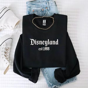 Puede incluir: Sudadera negra con el texto blanco "Disneyland est 1955". La imagen incluye vaqueros azul claro, un collar de cadena dorada y zapatillas altas blancas, sobre una superficie blanca.