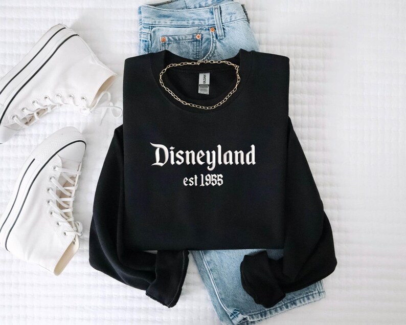 Puede incluir: Sudadera negra con el texto blanco "Disneyland est 1955". La imagen incluye vaqueros azul claro, un collar de cadena dorada y zapatillas altas blancas, sobre una superficie blanca.