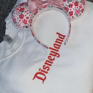 Puede incluir: Sudadera blanca con la palabra "Disneyland" bordada en rojo. Una diadema con estampado floral con orejas de rat&oacute;n y un lazo rosa se coloca sobre la sudadera. Las orejas tienen un borde festoneado blanco.