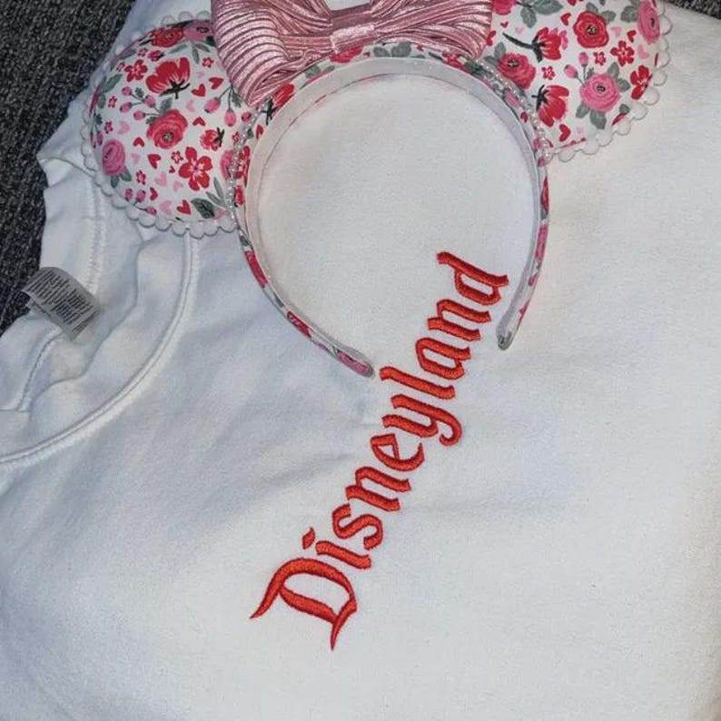 Puede incluir: Sudadera blanca con la palabra "Disneyland" bordada en rojo. Una diadema con estampado floral con orejas de rat&oacute;n y un lazo rosa se coloca sobre la sudadera. Las orejas tienen un borde festoneado blanco.