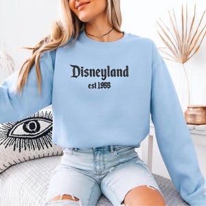 Puede incluir: Sudadera azul claro con el texto "Disneyland est 1955" en escritura negra. La parte superior de manga larga est&aacute; hecha de un material suave y es adecuada para ropa casual.