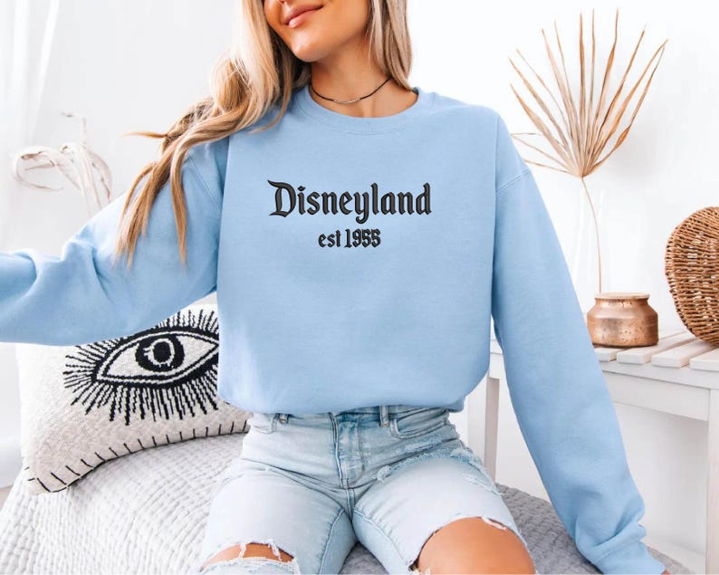 Puede incluir: Sudadera azul claro con el texto "Disneyland est 1955" en escritura negra. La parte superior de manga larga est&aacute; hecha de un material suave y es adecuada para ropa casual.