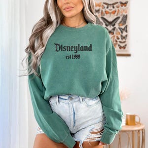 Puede incluir: Sudadera verde con cuello redondo con la palabra "Disneyland" en escritura negra y "est 1955" impresa debajo. La sudadera es de color verde liso y tiene mangas largas. La sudadera se combina con pantalones cortos de mezclilla.