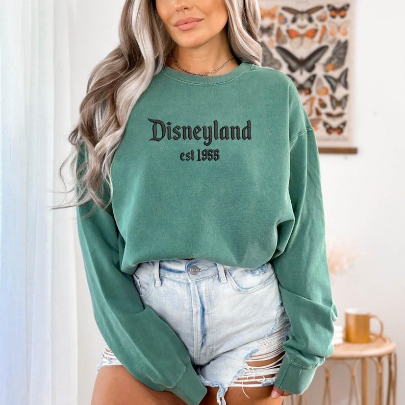 Puede incluir: Sudadera verde con cuello redondo con la palabra "Disneyland" en escritura negra y "est 1955" impresa debajo. La sudadera es de color verde liso y tiene mangas largas. La sudadera se combina con pantalones cortos de mezclilla.