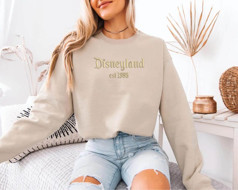 Puede incluir: Sudadera beige con la palabra "Disneyland" bordada en dorado, con "est 1955" debajo. La parte superior de manga larga es de color neutro, adecuada para ropa informal.