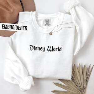 Puede incluir: Sudadera blanca bordada con el texto "Disney World" en fuente g&oacute;tica negra. Un collar dorado con un colgante est&aacute; sobre la camisa. La palabra "EMBROIDERED" est&aacute; impresa en una pancarta blanca.