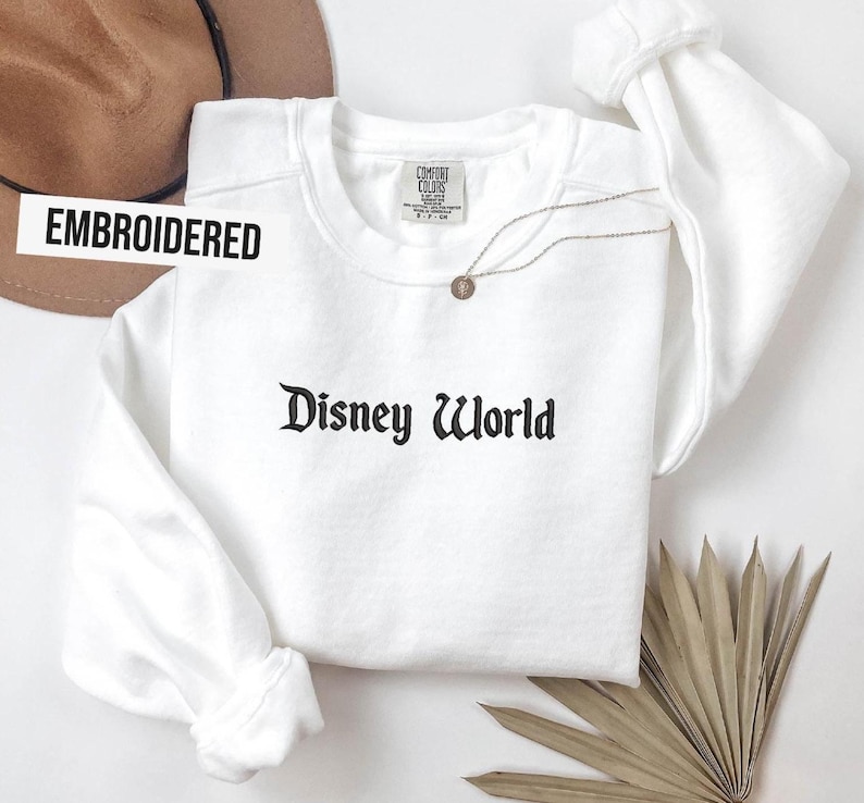 Puede incluir: Sudadera blanca bordada con el texto "Disney World" en fuente g&oacute;tica negra. Un collar dorado con un colgante est&aacute; sobre la camisa. La palabra "EMBROIDERED" est&aacute; impresa en una pancarta blanca.