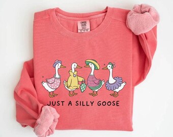 May include: Coral-colored sweatshirt featuring a whimsical illustration of four geese dressed in various outfits. The phrase "JUST A SILLY GOOSE" is printed below the geese. The sweatshirt has long sleeves with pink cuffs.
