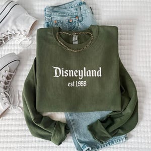 Puede incluir: Una sudadera verde oliva con la palabra "Disneyland" y el a&ntilde;o "est 1955" impresos en blanco. La sudadera se combina con vaqueros de mezclilla azul claro, zapatillas altas blancas y un collar de cadena dorada. Los art&iacute;culos est&aacute;n dispuestos sobre una superficie blanca texturizada.